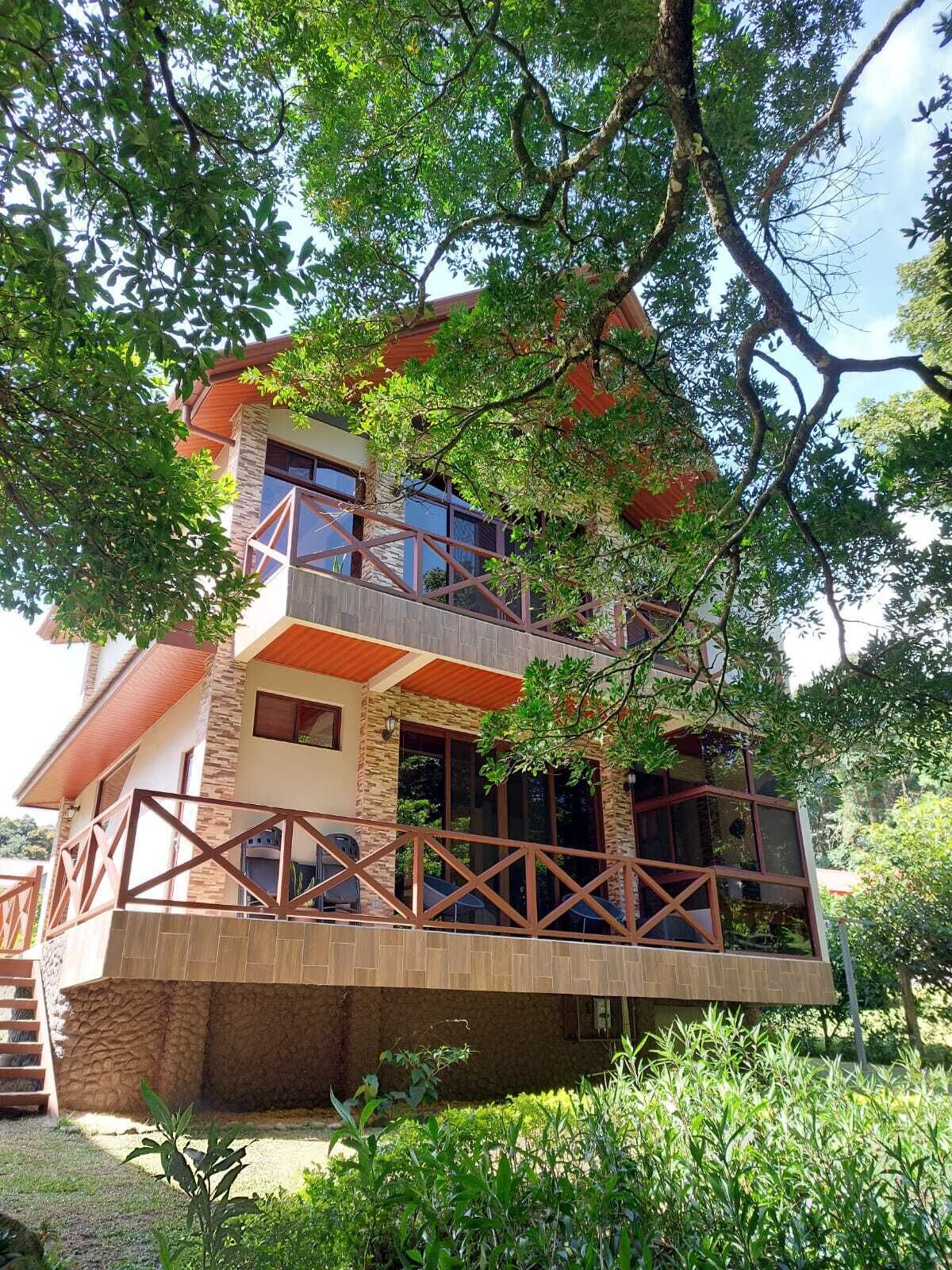 Una casa de dos pisos con un balcón, rodeada de árboles y follaje.