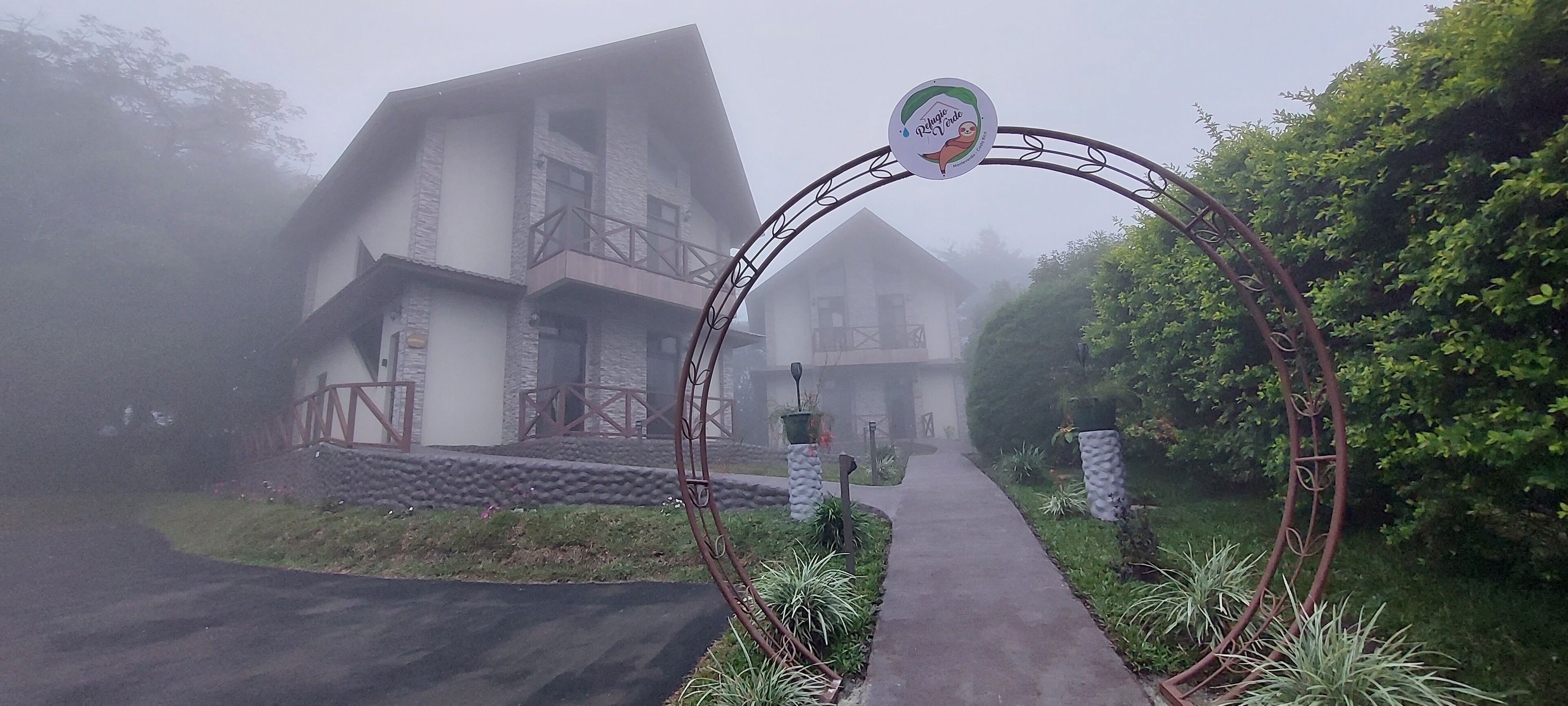 Un chemin menant à un bâtiment de style architectural rustique, entouré de verdure et surmonté d’une arche métallique portant un logo.