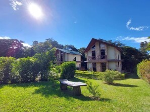 Una casa de dos pisos con fachada de piedra y un balcón, rodeada por un jardín cuidado y un cielo azul despejado.
