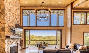 A spacious room with a stone fireplace, a chandelier, a leather sofa, and a large window offering a view of a deck and trees.