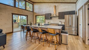 A modern kitchen with wooden floors, a central island with bar stools, stainless steel appliances, and large windows offering views of the outdoors. - EPIC Pool * View * Outdoor Kitchen * Luxury (Mineral Bluff)