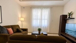 A modern living room with a brown sofa, a small table with a plant, a TV on a stand, and a window with sheer curtains. - FUN GAME St. Julians Apartment 3 (San Ġiljan)