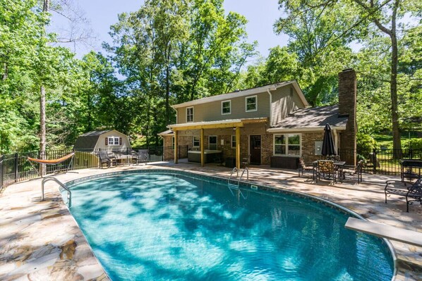 A swimming pool with a covered patio area, a stone fireplace, and a house with a porch.