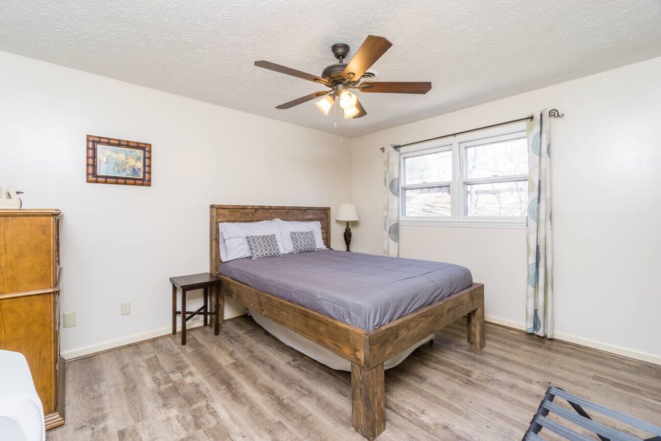 Un dormitorio con cama de madera, ventilador de techo, ventana con cortinas y una mesita de noche pequeña.