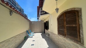 A narrow outdoor corridor with a tiled floor, a wooden door, and a small seating area with a table and chairs. - Modern House in Dunas Beach (cabo frio)