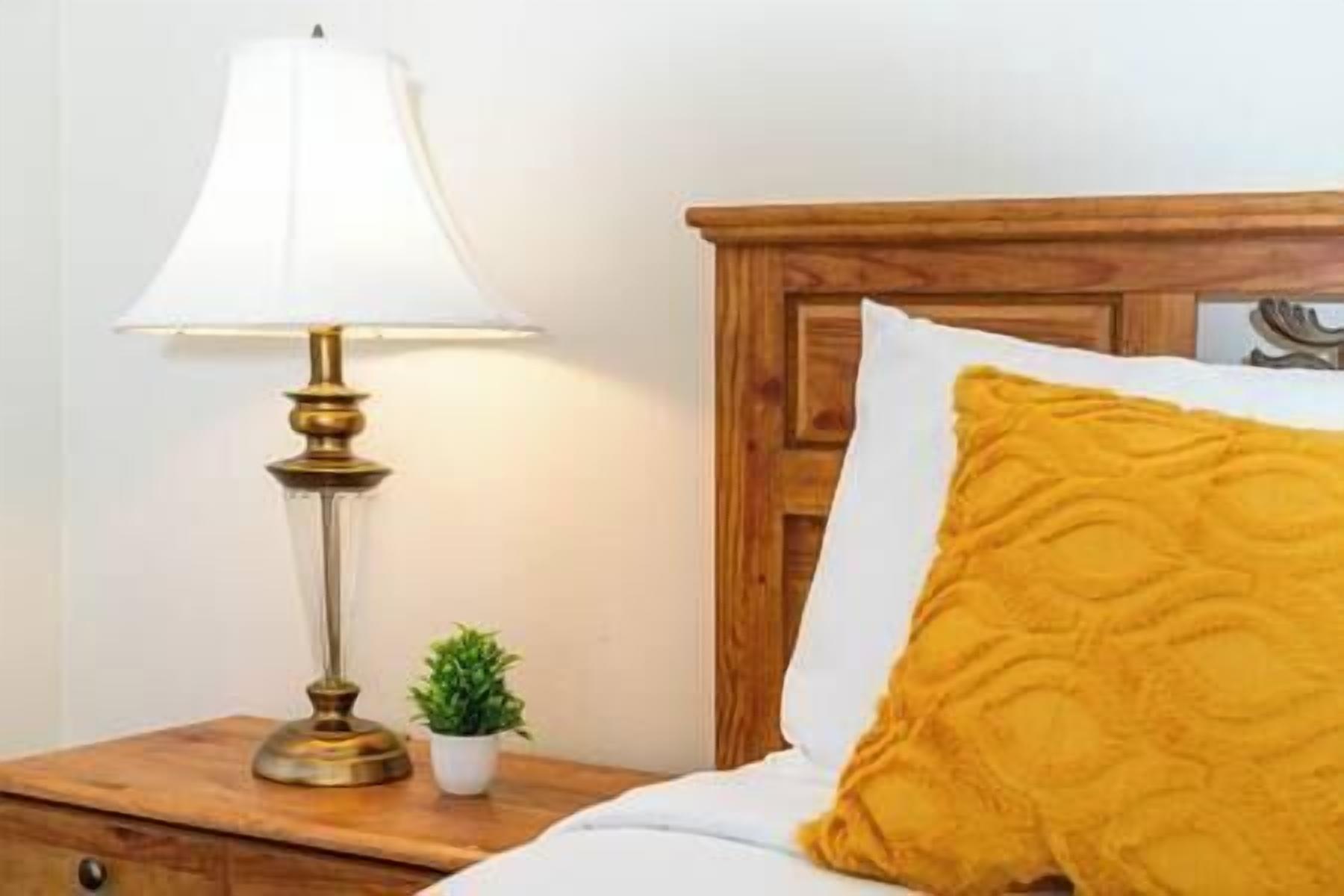 A bed with a wooden headboard, a white pillow, and a mustard-colored throw pillow. A small potted plant is on the bedside table next to a lamp with a white shade.