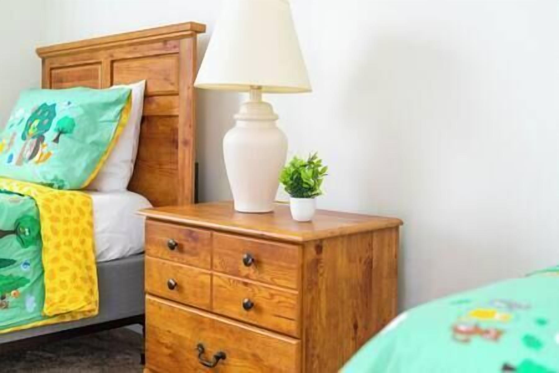 Een slaapkamer met een houten nachtkastje, een lamp, een kleine bloempot en een bed met groene beddengoed.