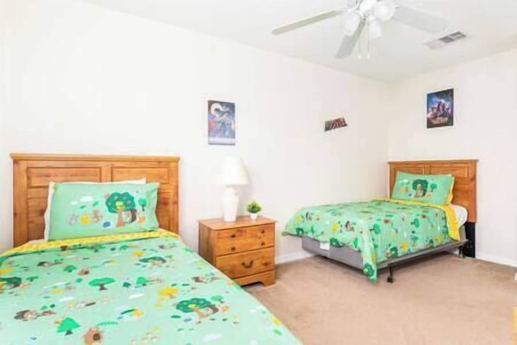 A bedroom with two beds, each with a green bedspread featuring cartoon characters. There is a wooden nightstand with a lamp and a small plant between the beds. The room has white walls and a ceiling fan.