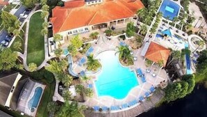 Eine Vogelperspektive zeigt ein Resort mit einem zentralen Schwimmbecken, umgeben von Liegebereichen und Palmen.