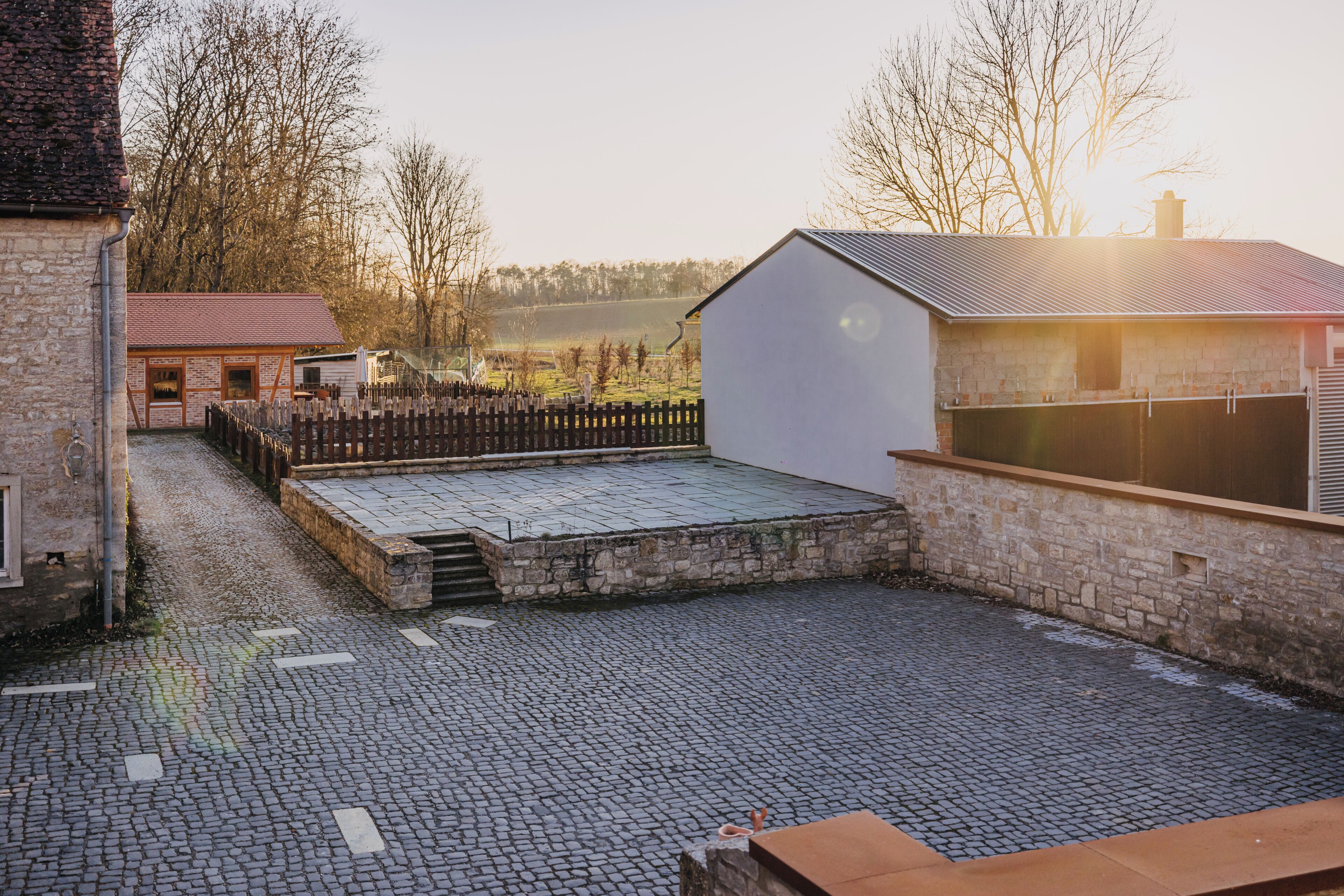 A courtyard with cobblestone flooring, a wooden fence, and a stone building with a red roof.