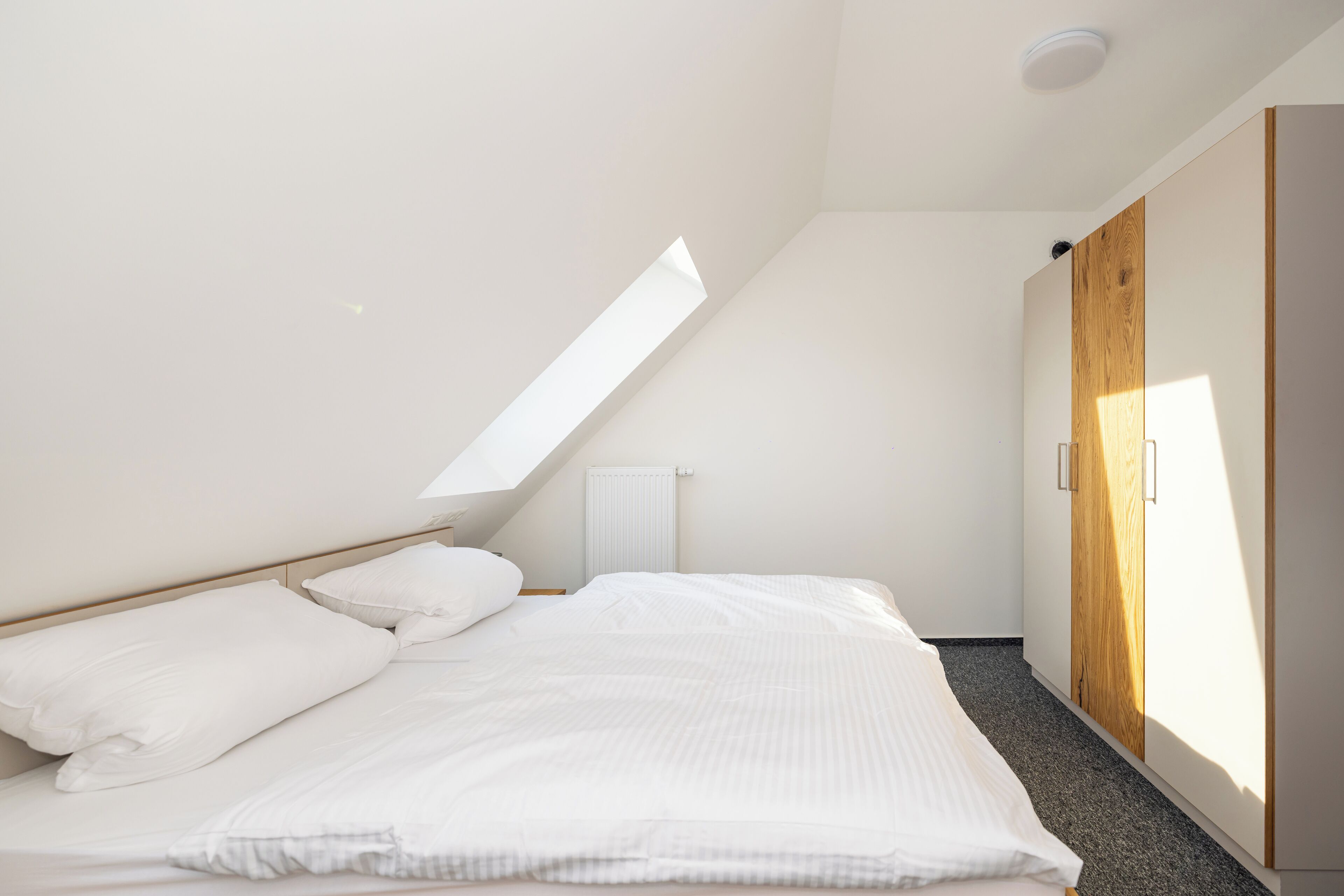 A bedroom with a sloped ceiling, a bed with white bedding, a wardrobe, and a radiator.