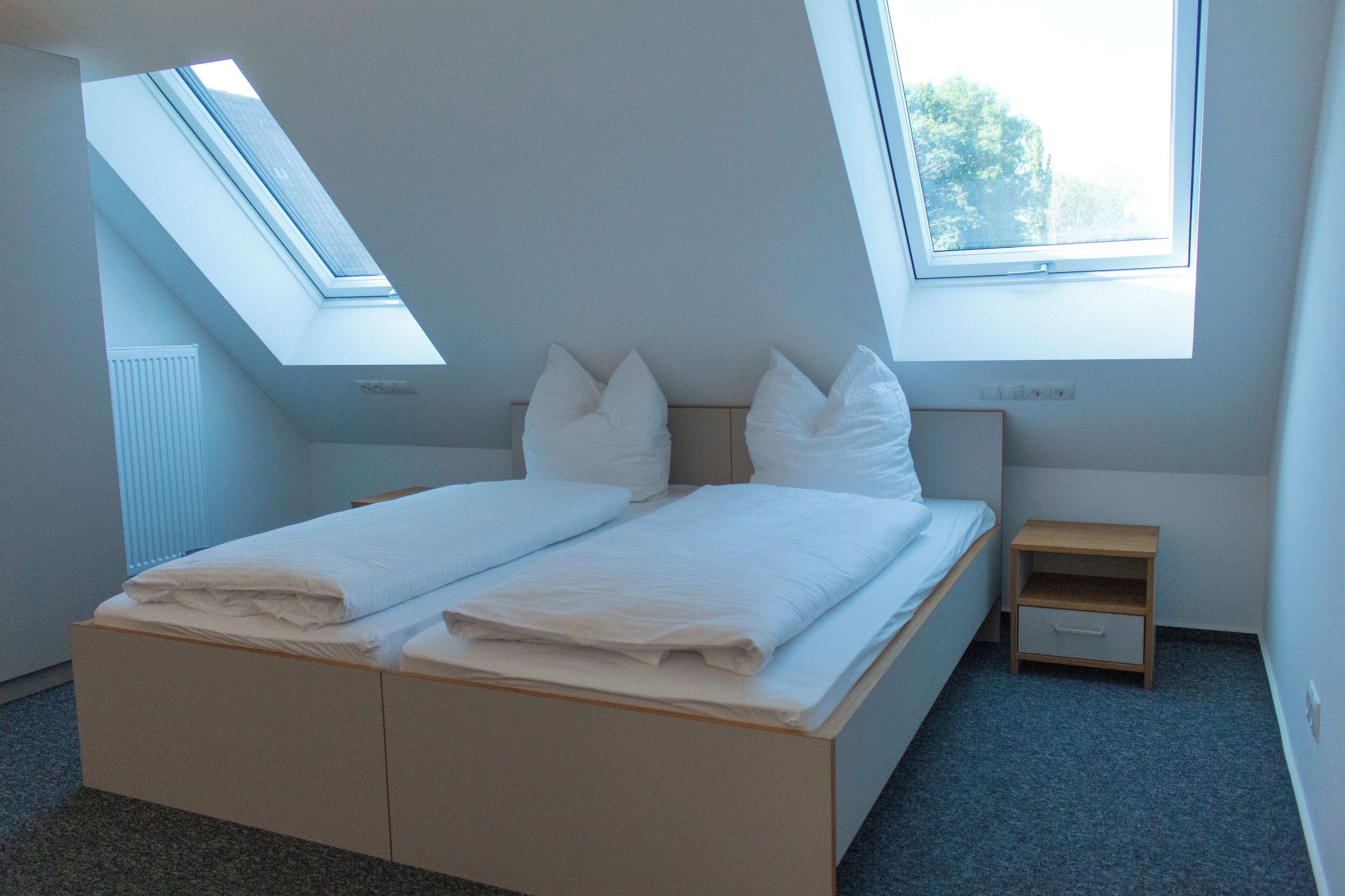 Un dormitorio con tragaluz, cama con ropa de cama blanca, mesita de noche y un mueble pequeño con cajones.