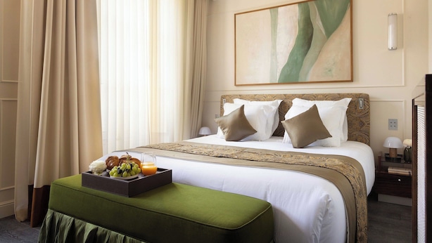 Une chambre d’hôtel avec un grand lit, une table de chevet, un plateau avec des fruits et du jus, et un pouf vert.