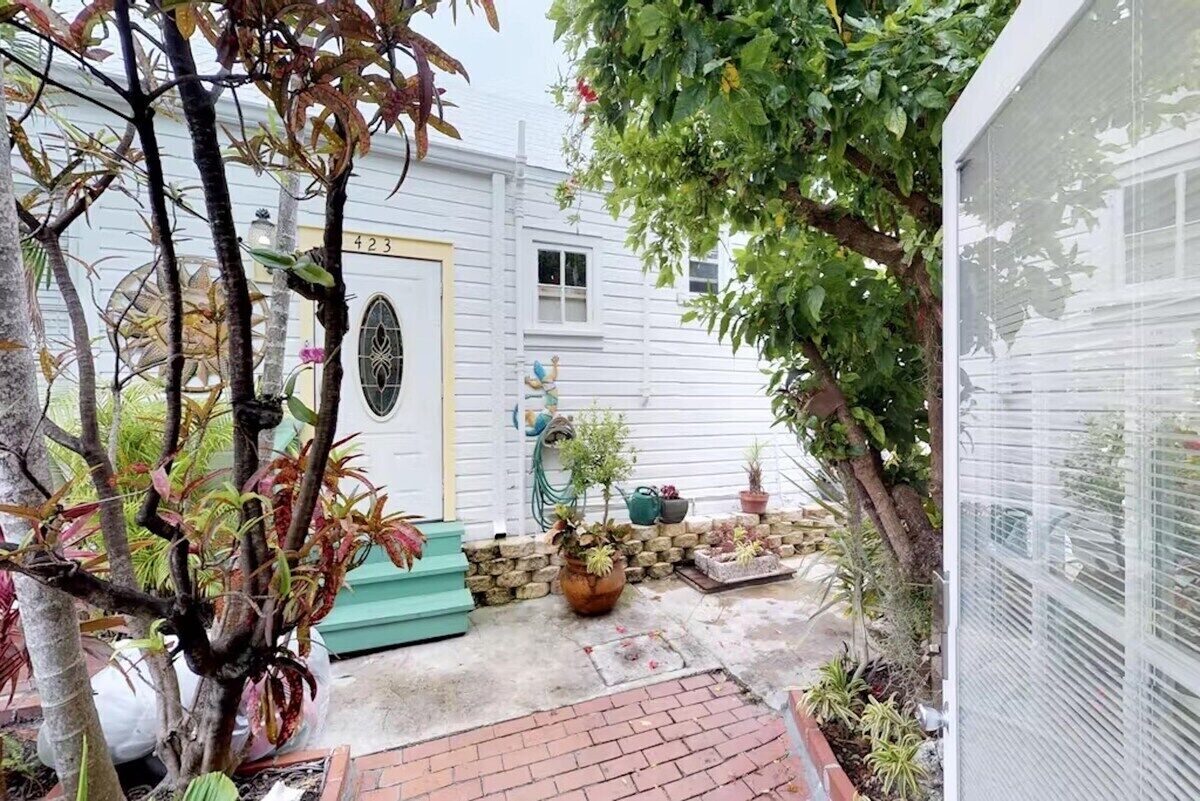 Un petit jardin clos, avec une maison blanche, une porte jaune et diverses plantes en pot.