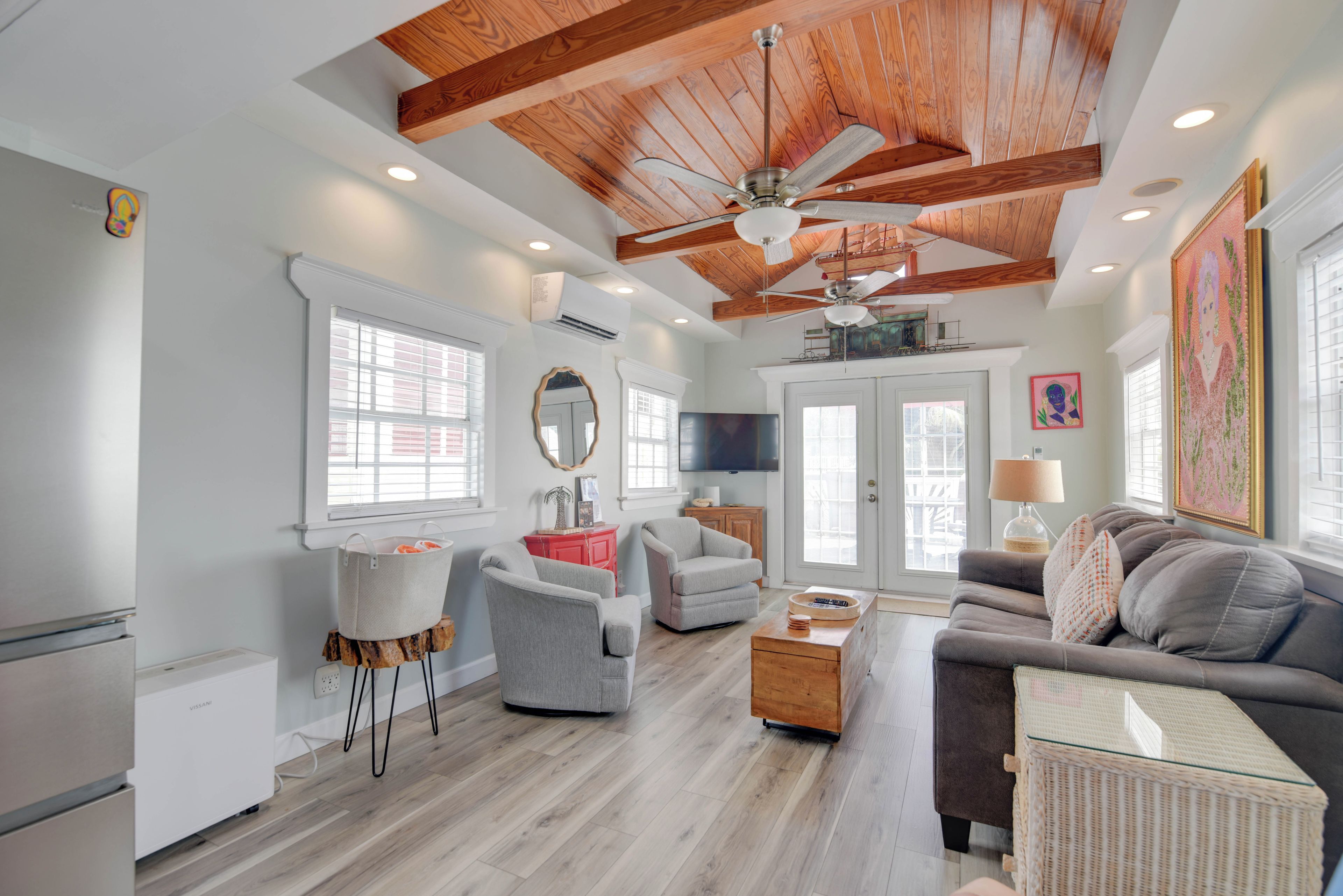 A spacious living room with a high ceiling, wooden beams, a ceiling fan, and a large window providing natural light.