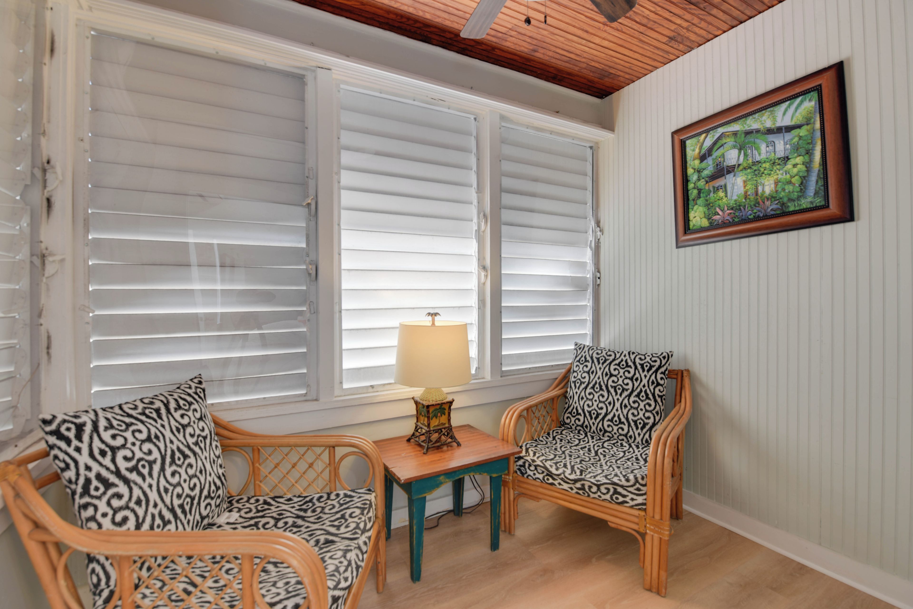 A room with two wicker chairs, a small table with a lamp, and a window with white shutters.