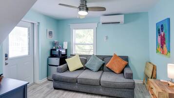 Un salon avec un canapé gris, un ventilateur de plafond, un climatiseur, une petite table et une fenêtre avec des stores.