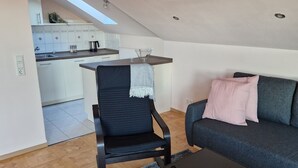 Una sala moderna con sofá, una butaca y una mesa de centro. Se observa un área de cocina al fondo.