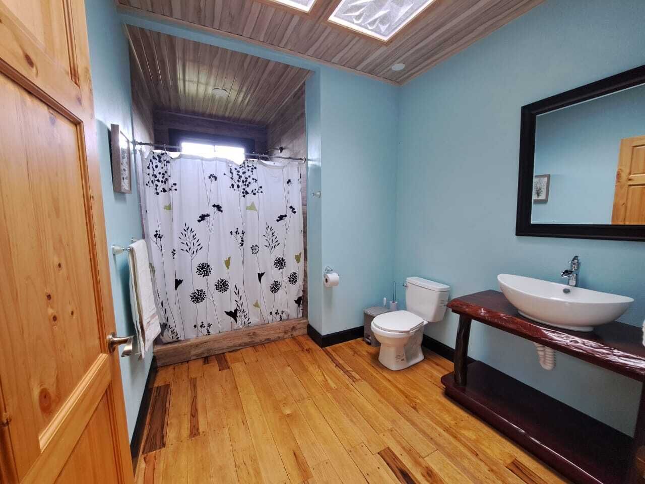 Ein Badezimmer mit Holzboden, einer weißen Toilette, einem hölzernen Waschtisch mit weißem Waschbecken und einer Duschvorhang mit floralem Muster.