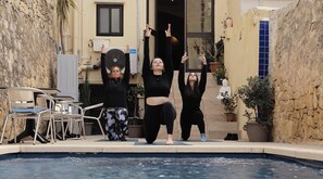 Three people performing yoga poses by a poolside with a stone wall and potted plants in the background. - Soul Dive World (Sliema)