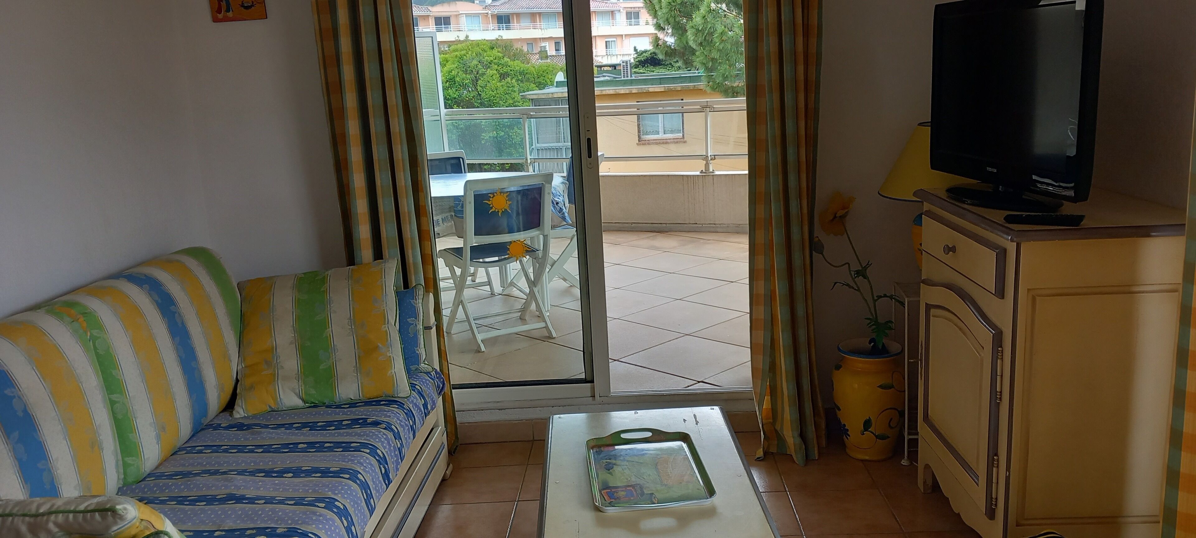 Un salon comprenant un canapé, une table basse, un téléviseur et un balcon offrant une vue sur les immeubles et la verdure.