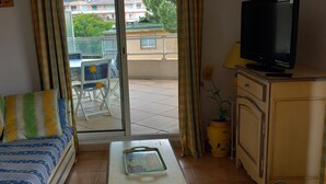 Un salon comprenant un canapé, une table basse, un téléviseur et un balcon offrant une vue sur les immeubles et la verdure.