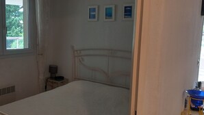 Une chambre avec un lit simple, une fenêtre, une table de chevet avec une lampe et un petit tabouret.