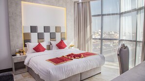 A modern hotel room with a large bed, a nightstand with a lamp, and a view of the city through large windows.