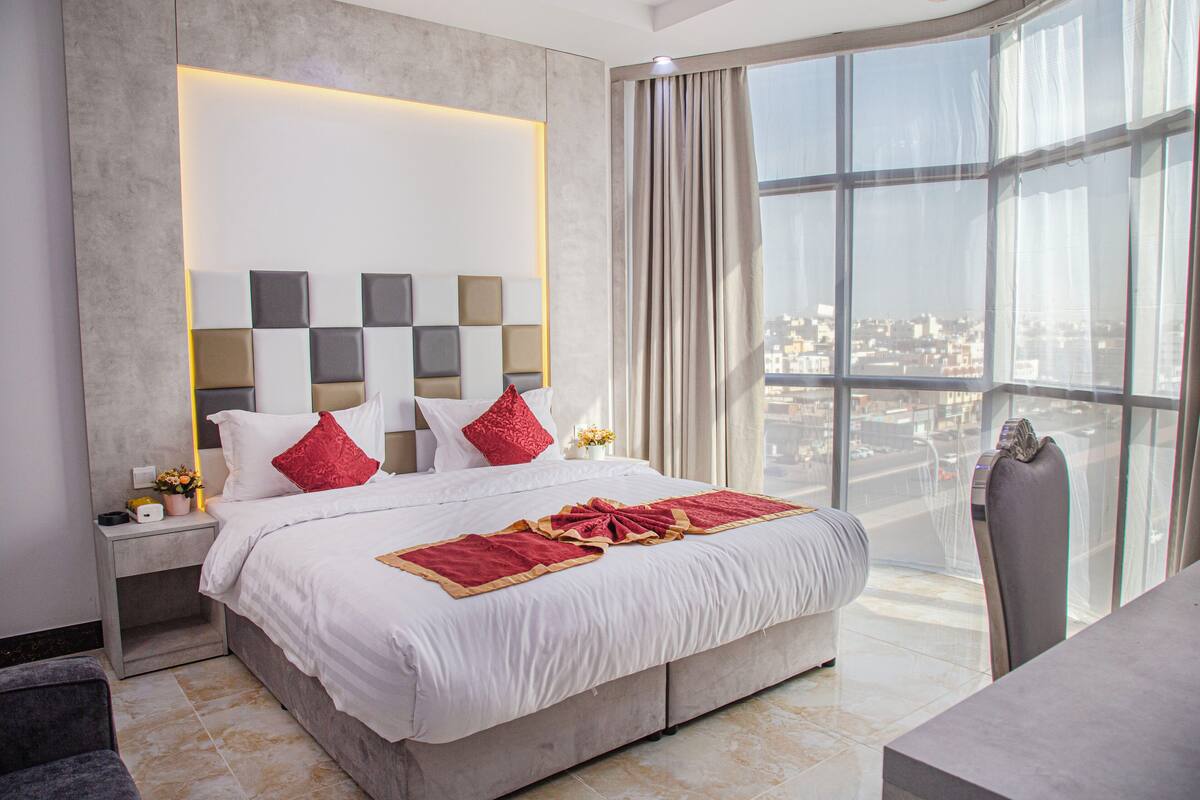 Een moderne hotelkamer met een groot bed, een nachtkastje met een lamp en uitzicht op de stad door grote ramen.