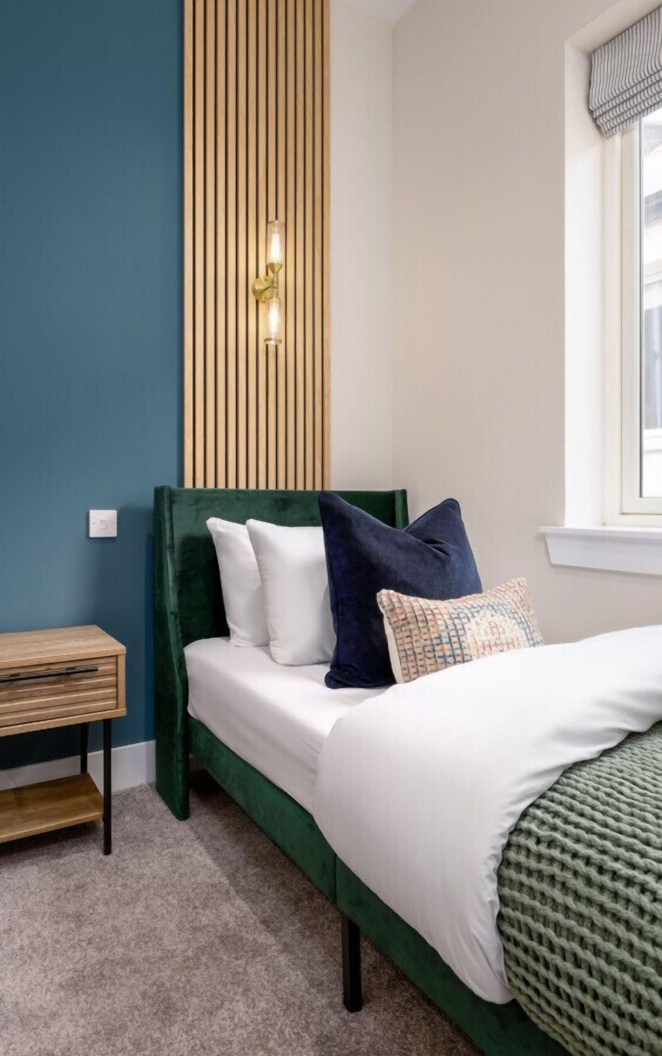 Un dormitorio ordenado con una cama tapizada en verde, almohadas blancas y con estampado, una mesita de noche de madera y una lámpara de pared.