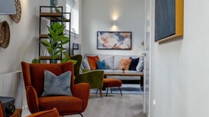A modern living room with a sofa, armchairs, and a coffee table. There is a plant, a painting on the wall, and a window providing natural light.