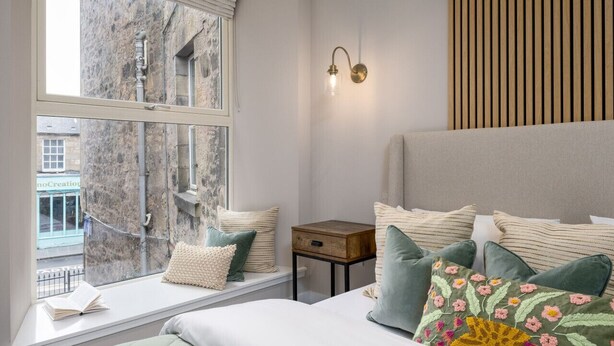 Een slaapkamer met een groot bed, een nachtkastje, een raam met uitzicht op gebouwen, en een wandlamp.