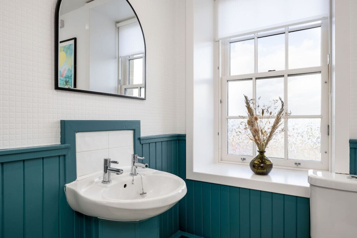 Une salle de bain avec un lavabo blanc, un lambris mural de couleur turquoise, une fenêtre avec vue et un miroir au-dessus du lavabo.