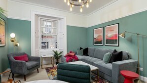 Ein Wohnzimmer mit einem grauen Sofa, einem grünen Sessel, einem roten Beistelltisch und einem weißen Couchtisch. Ein Fenster bietet Blick auf einen Lagerraum, auf der Fensterbank steht eine Pflanze.