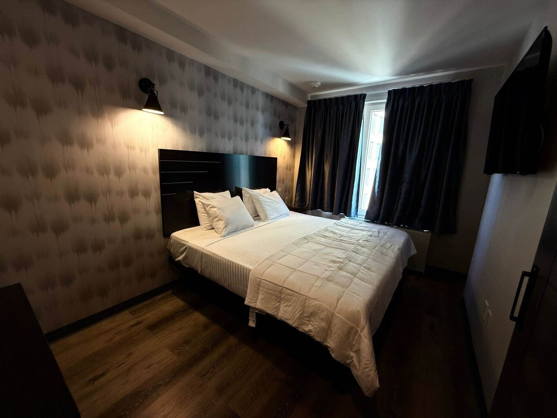 Een hotelkamer met een groot bed, donkere gordijnen, een wandtelevisie en een houten vloer.