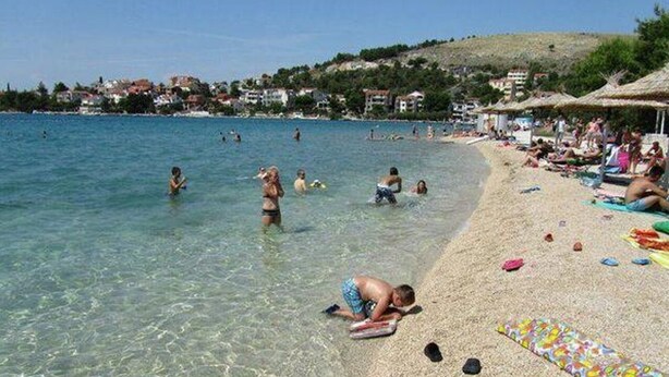 Una playa con agua cristalina, gente nadando y tomando el sol, y una hilera de cabañas de playa al fondo.