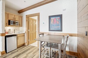A compact kitchen with wooden cabinets, a microwave, and a sink. There's a dining table with chairs and a framed picture on the wall.