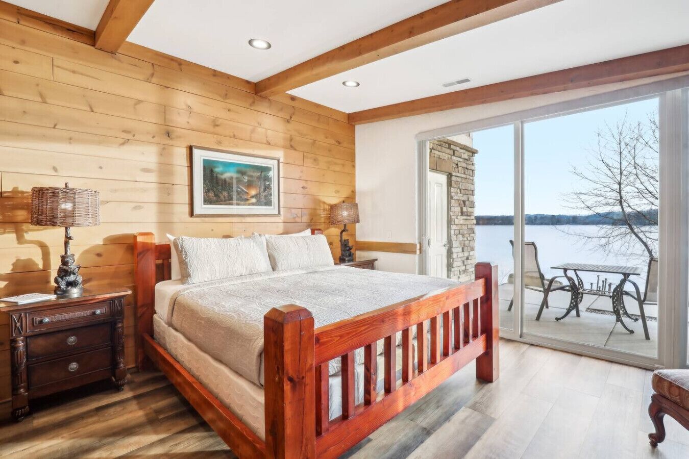 A wooden bedroom with a large bed, wooden nightstands, a table by the window, and a view of a lake.