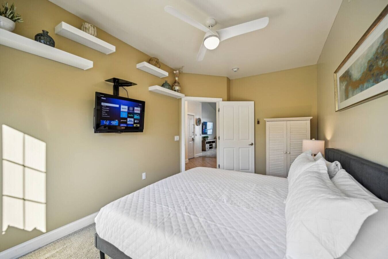 Une chambre à coucher avec un lit, un ventilateur de plafond, un téléviseur fixé au mur et une porte donnant sur une autre pièce.