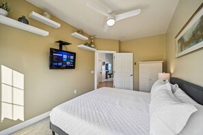 Une chambre à coucher avec un lit, un ventilateur de plafond, un téléviseur fixé au mur et une porte donnant sur une autre pièce.