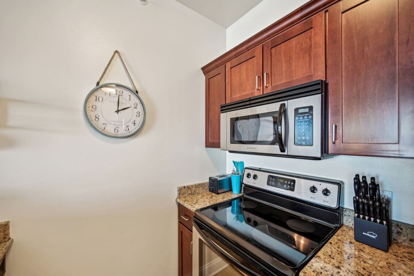 A kitchen with wooden cabinets, a stainless steel microwave, an oven, and a wall clock.