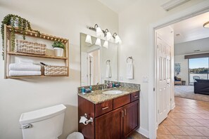 Un baño con inodoro blanco, tocador de madera con encimera de granito y un espejo grande con luces empotradas.