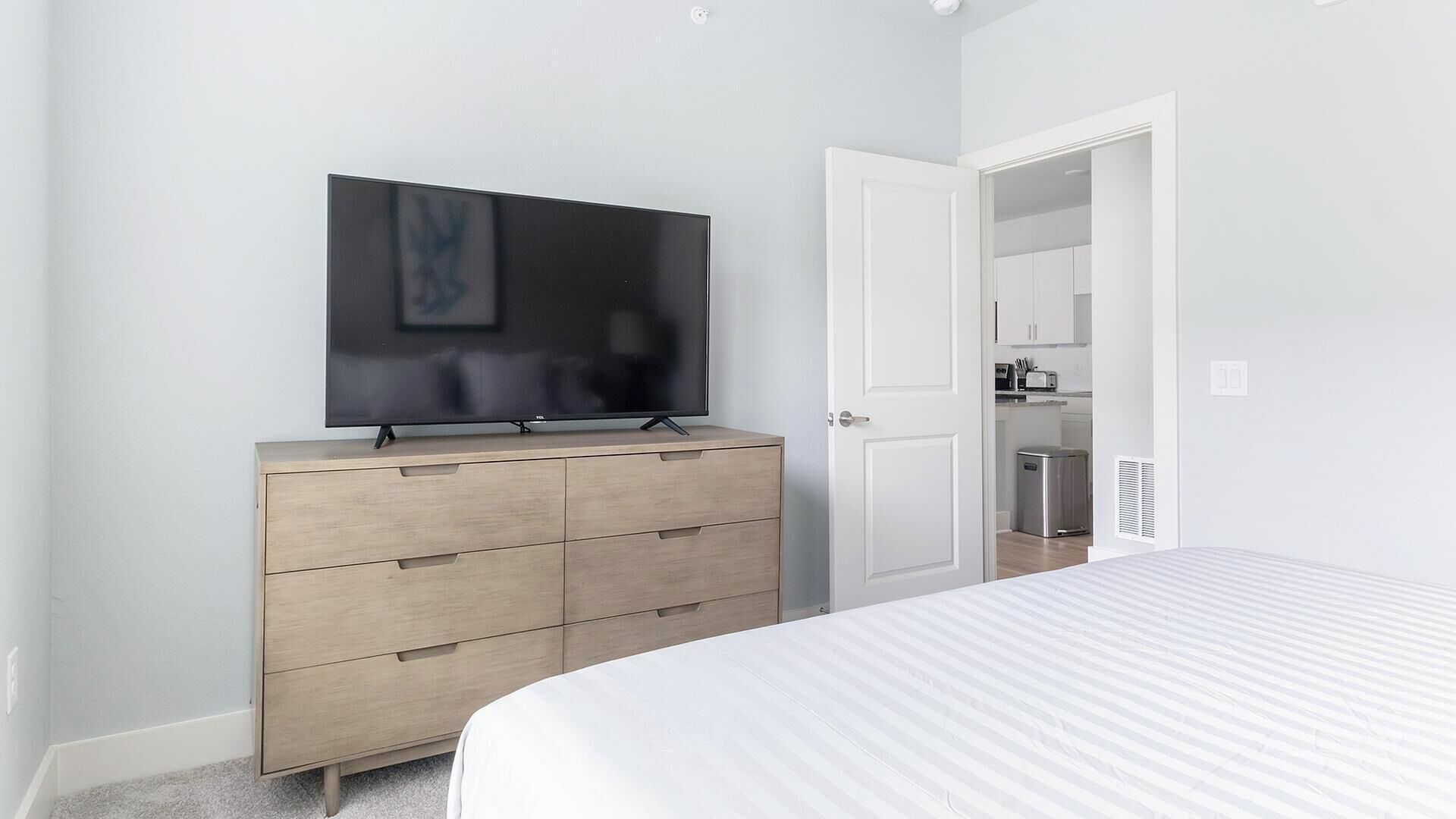 A bedroom with a bed, a dresser with a flat-screen TV, and a door leading to a kitchenette.
