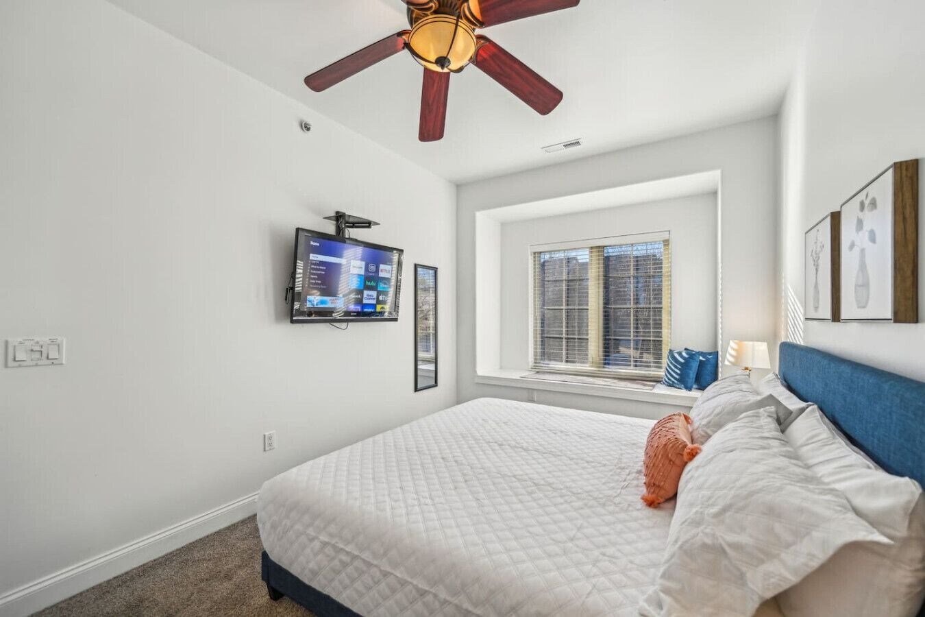 Ein Schlafzimmer mit einem Bett, einem Deckenventilator, einem an der Wand montierten Fernseher und einem Fenster mit Jalousien.