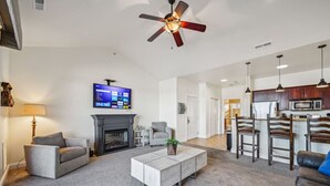 Un salon doté d’une cheminée, d’un ventilateur de plafond, d’un téléviseur à écran plat et d’un coin cuisine avec des tabourets de bar.