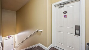 Un couloir aux murs de couleur beige, une porte blanche avec une plaque numérotée et un garde-corps menant à une sortie.