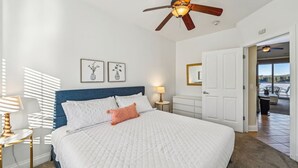 Une chambre à coucher comprenant un lit, un ventilateur de plafond, une table de chevet, une lampe, une commode et une porte donnant sur un balcon.