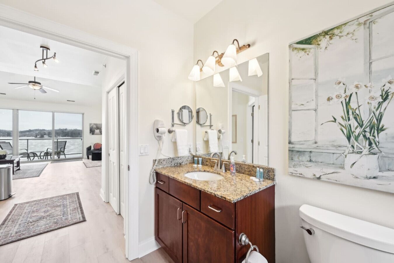 A bathroom with a granite countertop, a toilet, a large mirror, and a view of a balcony with a chair and a table.