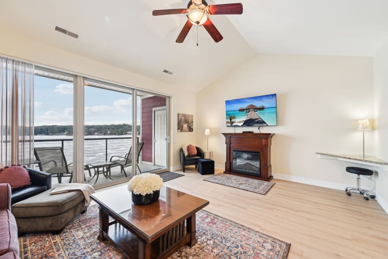 A spacious living room with a large window, a fireplace, a ceiling fan, and a television mounted on the wall.