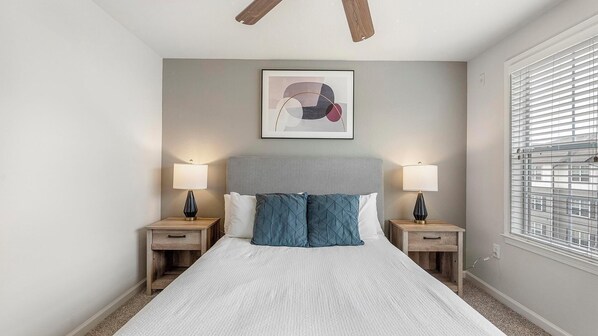 Un dormitorio con cama, dos mesitas de noche, dos lámparas, un ventilador de techo, una obra de arte enmarcada y una ventana con persianas.
